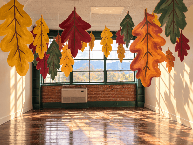 An empty studio room with polished hardwood floors and large grid windows, decorated with numerous oversized cardboard autumn leaves in shades of red, yellow, orange, and green hanging suspended from the ceiling.